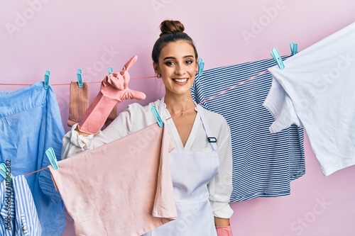 Beautiful brunette young woman washing clothes at clothesline smiling and confident gesturing with hand doing small size sign with fingers looking and the camera. measure concept.
