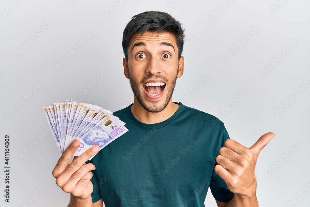 Young handsome man holding 20 swedish krona banknotes pointing thumb up to the side smiling happy with open mouth Young handsome man holding 20 swedish krona banknotes pointing thumb up to the side smiling happy with open mouth