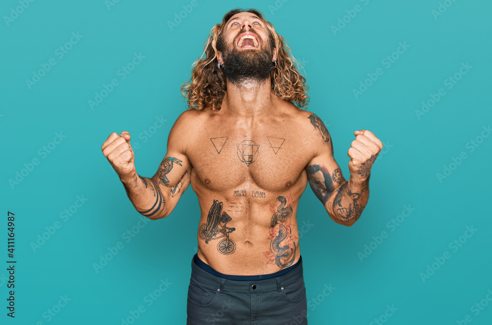 Obraz premium Handsome man with beard and long hair standing shirtless showing tattoos crazy and mad shouting and yelling with aggressive expression and arms raised. frustration concept.