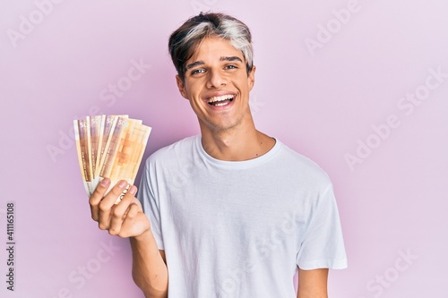 Young hispanic man holding 500 norwegian krone banknotes looking positive and happy standing and smiling with a confident smile showing teeth photo