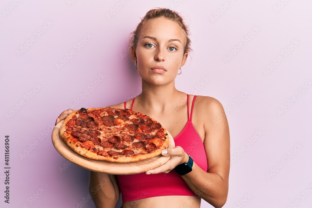 Beautiful caucasian woman holding italian pizza relaxed with serious expression on face. simple and natural looking at the camera.