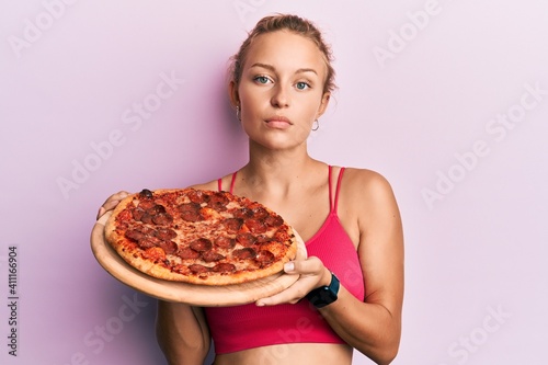 Beautiful caucasian woman holding italian pizza relaxed with serious expression on face. simple and natural looking at the camera.