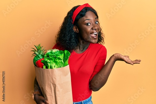 Beautiful african young woman holding paper bag with groceries celebrating achievement with happy smile and winner expression with raised hand photo