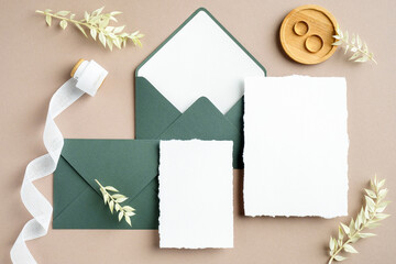 Elegant wedding stationery set. Wedding invitation cards templates, green envelopes, silk ribbon, golden rings on pastel beige background. Flat lay, top view, copy space.