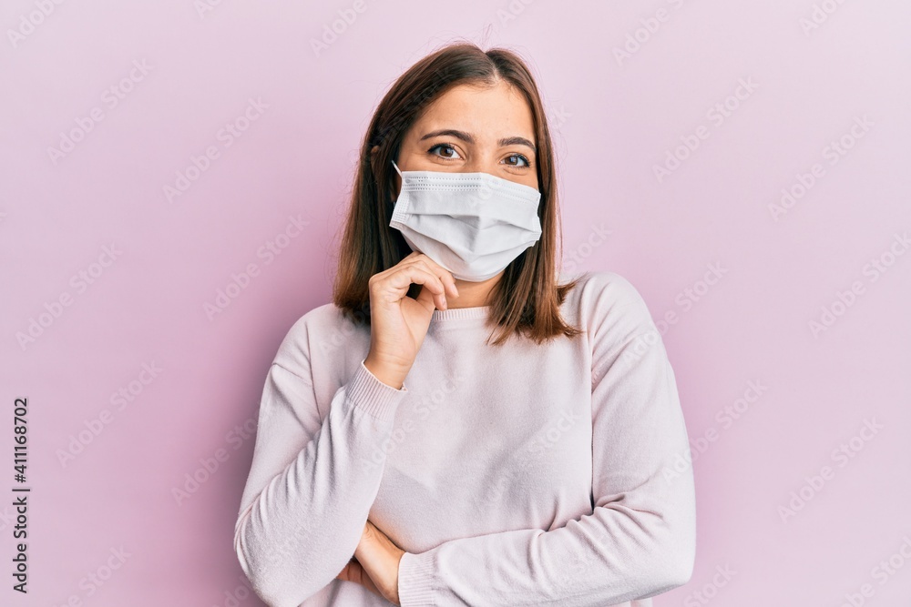 Young beautiful woman wearing medical mask smiling looking confident at the camera with crossed arms and hand on chin. thinking positive. Young beautiful woman wearing medical mask smiling looking confident at the camera with crossed arms and hand on chin. thinking positive.