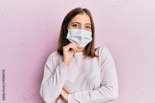 Young beautiful woman wearing medical mask smiling looking confident at the camera with crossed arms and hand on chin. thinking positive.