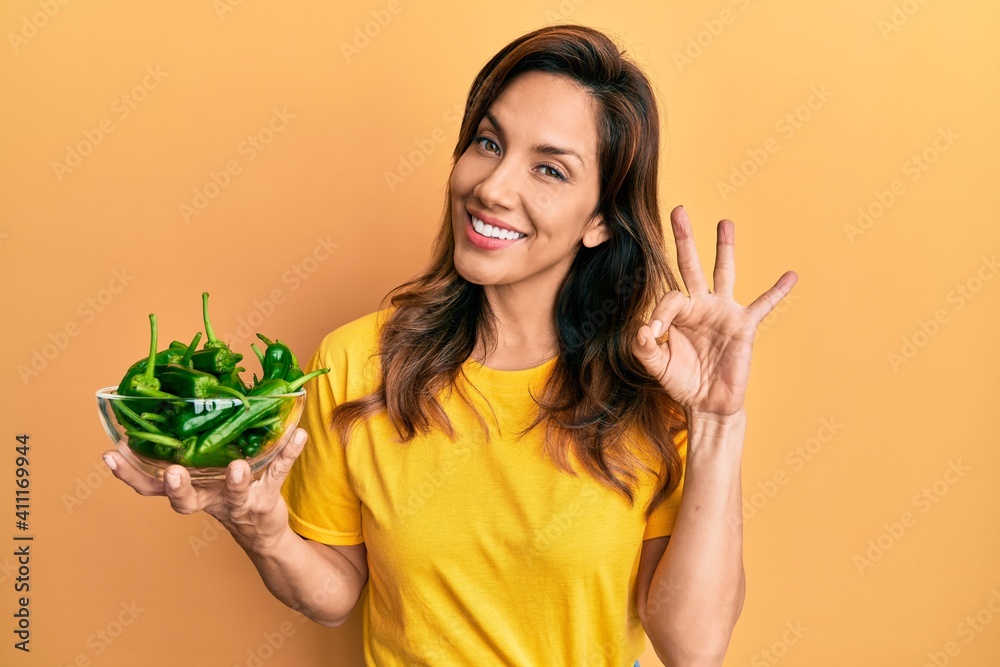 Young latin woman holding bowl with green peppers doing ok sign with fingers, smiling friendly gesturing excellent symbol Young latin woman holding bowl with green peppers doing ok sign with fingers, smiling friendly gesturing excellent symbol