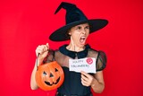 Young hispanic woman wearing witch costume holding pumpkin and happy halloween message angry and mad screaming frustrated and furious, shouting with anger. rage and aggressive concept.