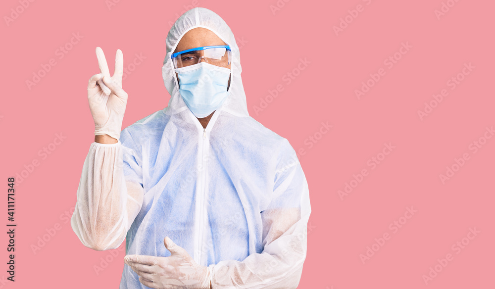 Young hispanic man wearing doctor protection coronavirus uniform and medical mask smiling with happy face winking at the camera doing victory sign. number two. Young hispanic man wearing doctor protection coronavirus uniform and medical mask smiling with happy face winking at the camera doing victory sign. number two.