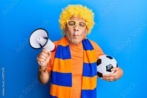 Senior hispanic man football hooligan cheering game holding ball and megaphone making fish face with mouth and squinting eyes, crazy and comical.