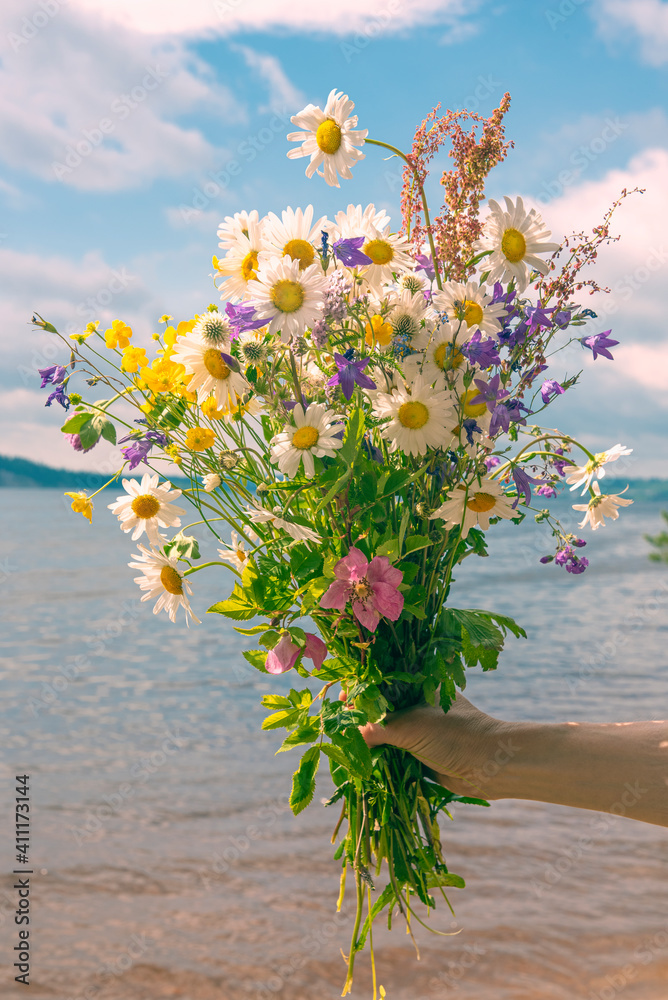 A bouquet of wild flowers in your hand on the background of the river and the sky with clouds.