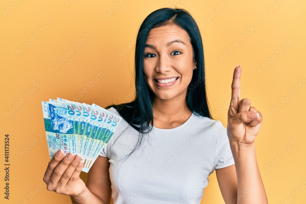 Beautiful hispanic woman holding hong kong 20 dollars banknotes smiling with an idea or question pointing finger with happy face, number one Beautiful hispanic woman holding hong kong 20 dollars banknotes smiling with an idea or question pointing finger with happy face, number one