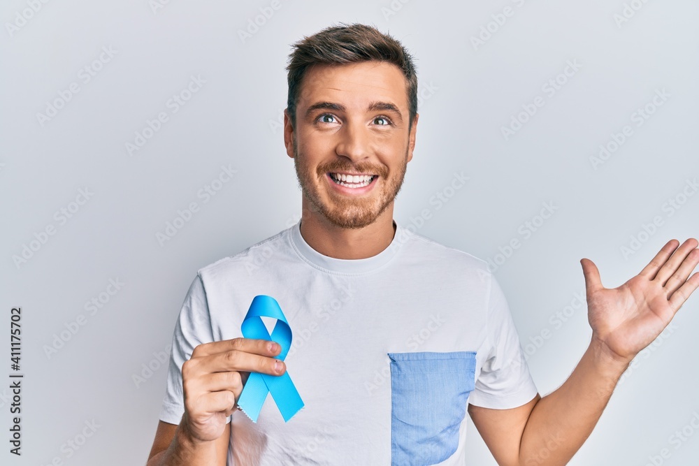 Handsome caucasian man holding blue ribbon celebrating victory with happy smile and winner expression with raised hands Handsome caucasian man holding blue ribbon celebrating victory with happy smile and winner expression with raised hands