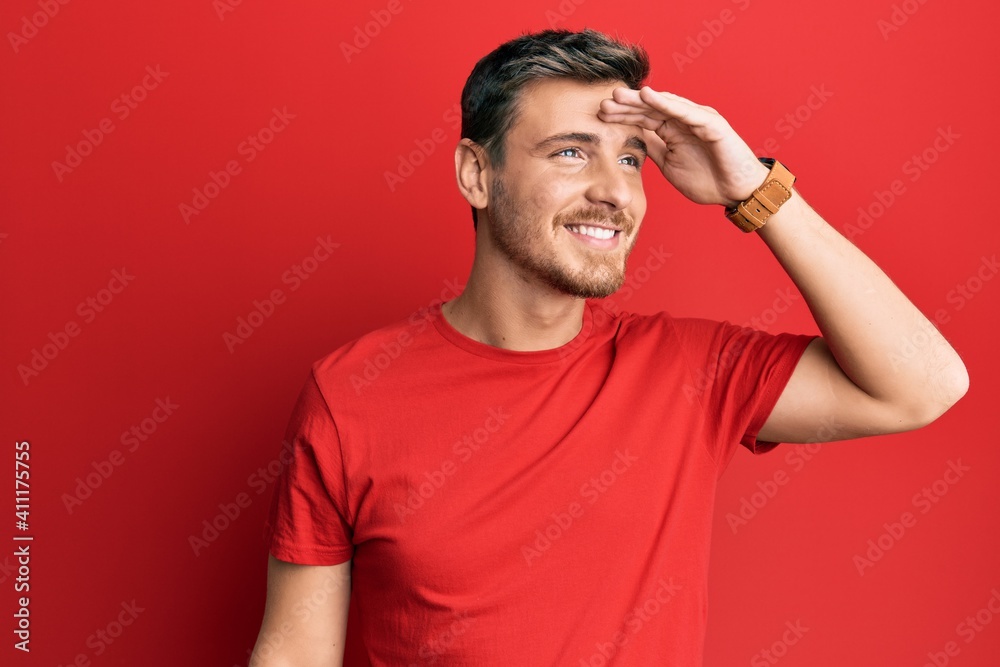 Handsome caucasian man wearing casual red tshirt very happy and smiling looking far away with hand over head. searching concept. Handsome caucasian man wearing casual red tshirt very happy and smiling looking far away with hand over head. searching concept.