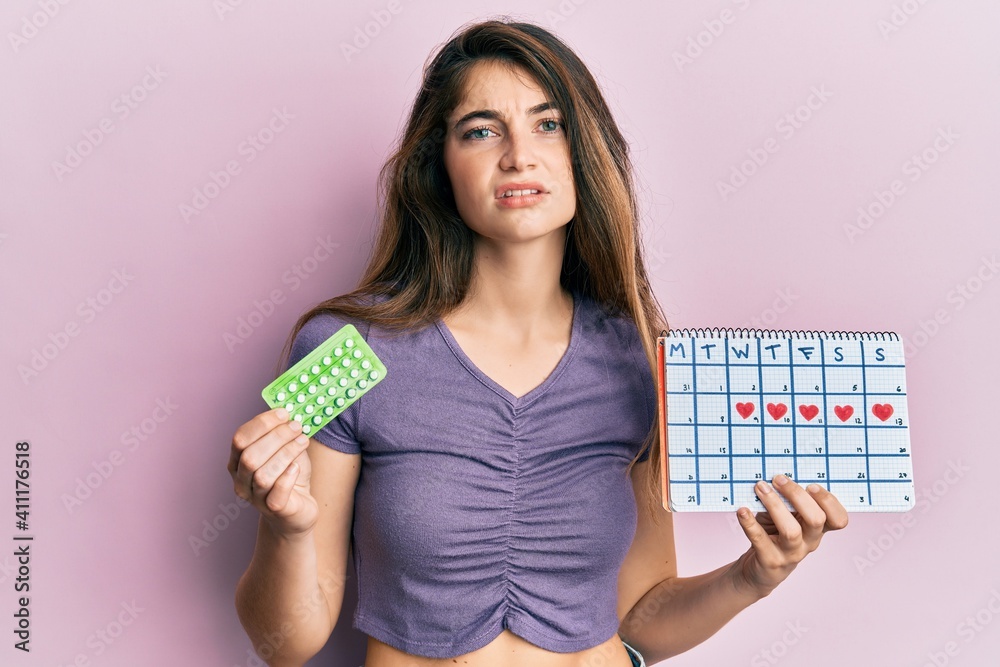 Young caucasian woman holding birth control pills and menstrual calendar clueless and confused expression. doubt concept. Young caucasian woman holding birth control pills and menstrual calendar clueless and confused expression. doubt concept.