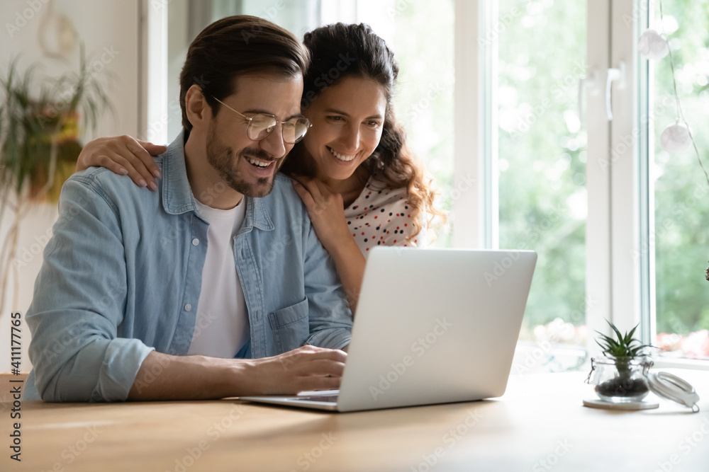 Cheering spouses having warm relations hug by desk at home make shopping purchases online at web store. Friendly young couple in love look at computer screen planning wedding choosing tour on vacation Cheering spouses having warm relations hug by desk at home make shopping purchases online at web store. Friendly young couple in love look at computer screen planning wedding choosing tour on vacation