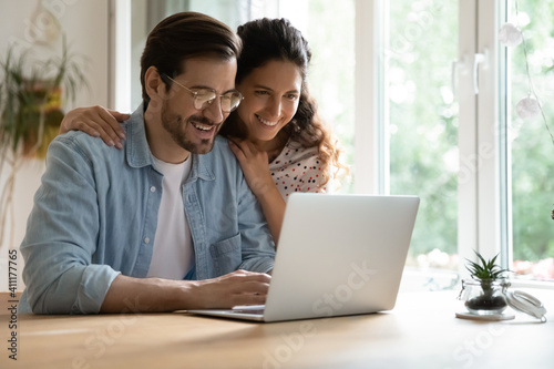 Cheering spouses having warm relations hug by desk at home make shopping purchases online at web store. Friendly young couple in love look at computer screen planning wedding choosing tour on vacation