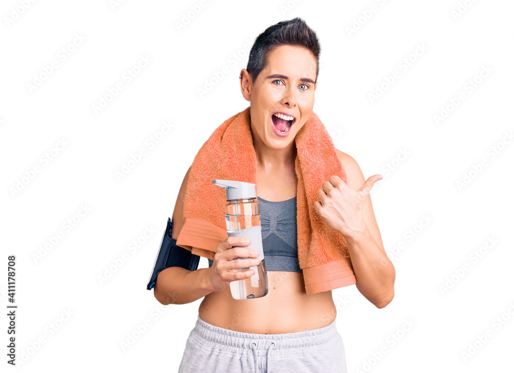 Young woman with short hair wearing sportswear and towel holding bottle of water pointing thumb up to the side smiling happy with open mouth Young woman with short hair wearing sportswear and towel holding bottle of water pointing thumb up to the side smiling happy with open mouth