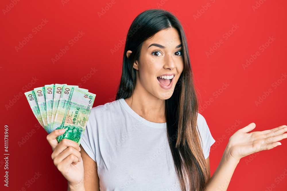Young brunette woman holding hong kong 50 dollars banknotes celebrating achievement with happy smile and winner expression with raised hand Young brunette woman holding hong kong 50 dollars banknotes celebrating achievement with happy smile and winner expression with raised hand