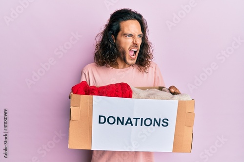 Young handsome man with long hair holding donations box for charity angry and mad screaming frustrated and furious, shouting with anger. rage and aggressive concept. © Krakenimages.com