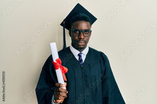 Handsome black man wearing graduation cap and ceremony robe holding diploma thinking attitude and sober expression looking self confident photo