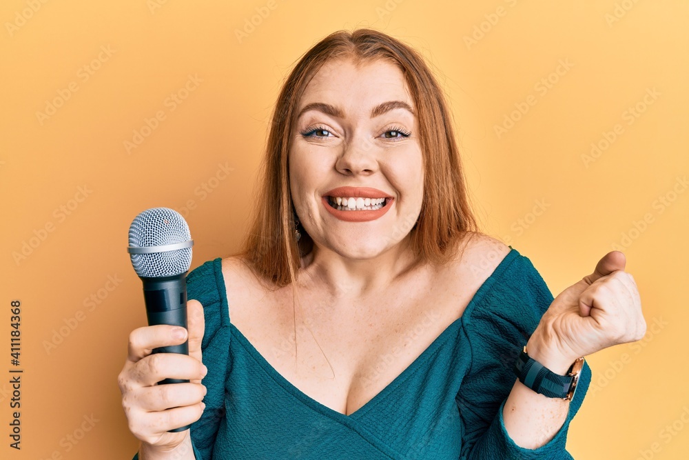 Young beautiful redhead woman singing song using microphone screaming proud, celebrating victory and success very excited with raised arm Young beautiful redhead woman singing song using microphone screaming proud, celebrating victory and success very excited with raised arm