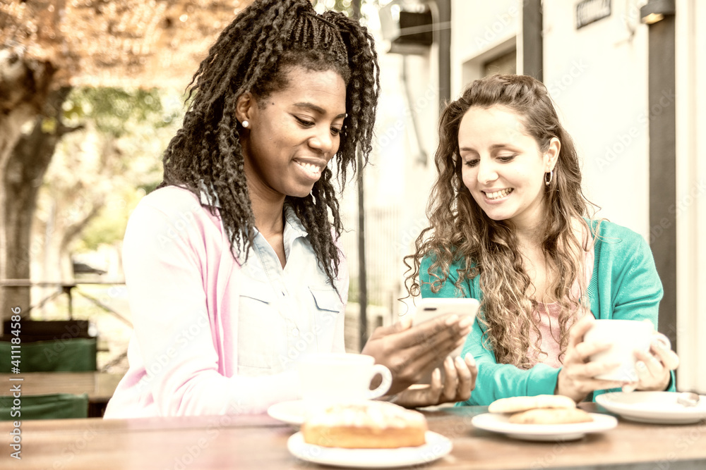 Two nice young woman watch mobile phone, drink coffee and eat cakes