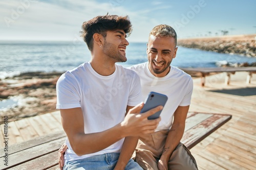 Young gay couple smiling happy using smartphone at the beach.