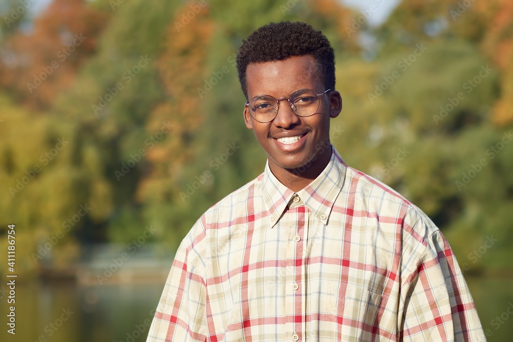 Portrait of handsome happy black African Afro American young man in shirt and glasses standing outdoors in a golden autumn park, smiling, looking at camera with intelligent smart face. Positive guy Portrait of handsome happy black African Afro American young man in shirt and glasses standing outdoors in a golden autumn park, smiling, looking at camera with intelligent smart face. Positive guy