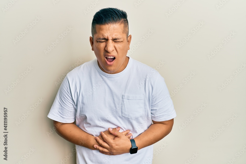 Young latin man wearing casual white t shirt with hand on stomach because nausea, painful disease feeling unwell. ache concept. Young latin man wearing casual white t shirt with hand on stomach because nausea, painful disease feeling unwell. ache concept.