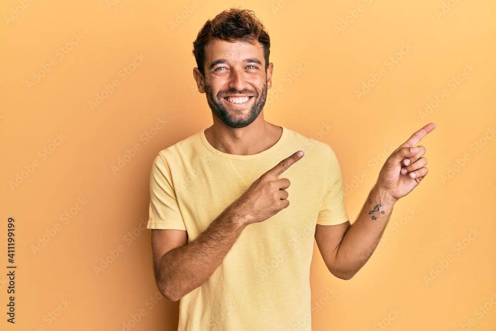 Handsome man with beard wearing casual yellow tshirt over yellow background smiling and looking at the camera pointing with two hands and fingers to the side.