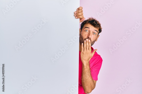 Handsome man with beard holding blank empty banner covering mouth with hand, shocked and afraid for mistake. surprised expression photo