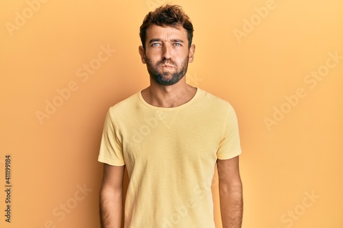 Handsome man with beard wearing casual yellow tshirt over yellow background puffing cheeks with funny face. mouth inflated with air, crazy expression.