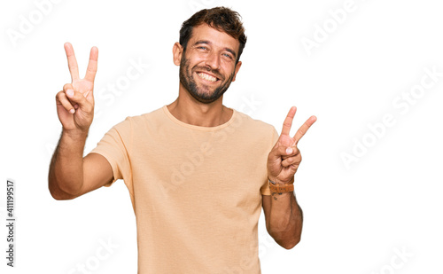 Handsome young man with beard wearing casual tshirt smiling with tongue out showing fingers of both hands doing victory sign. number two.