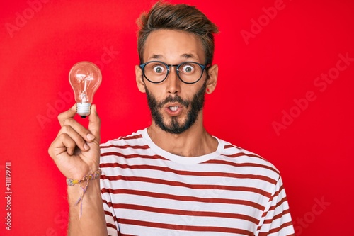 Handsome caucasian man with beard holding lightbulb for inspiration and idea scared and amazed with open mouth for surprise, disbelief face