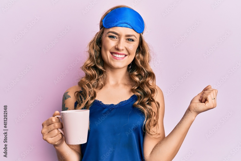 Young blonde girl wearing sleep mask and pyjama drinking coffee screaming proud, celebrating victory and success very excited with raised arm