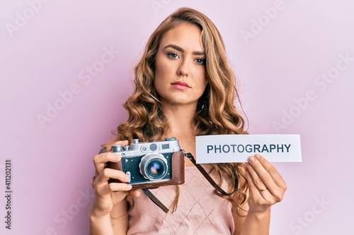 Young blonde girl holding vintage camera and paper with photography word paper relaxed with serious expression on face. simple and natural looking at the camera.