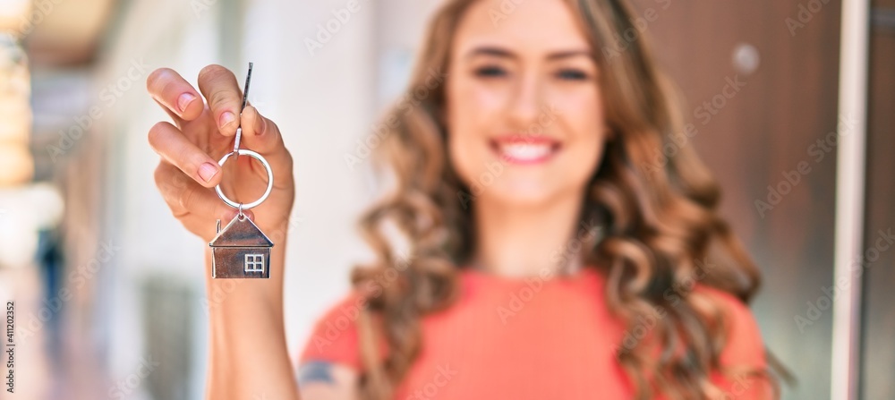 Young blonde girl smiling happy holding key of new house standing at the city. - obrazy, fototapety, plakaty 