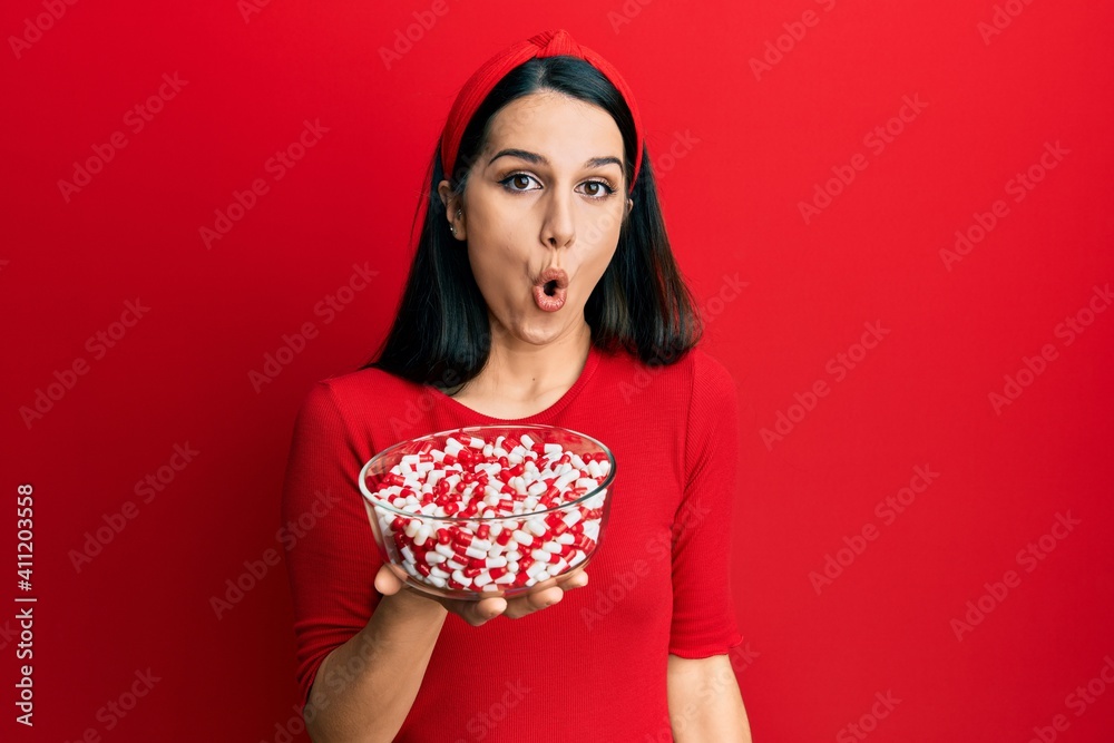Young hispanic woman holding bowl of pills scared and amazed with open mouth for surprise, disbelief face Young hispanic woman holding bowl of pills scared and amazed with open mouth for surprise, disbelief face