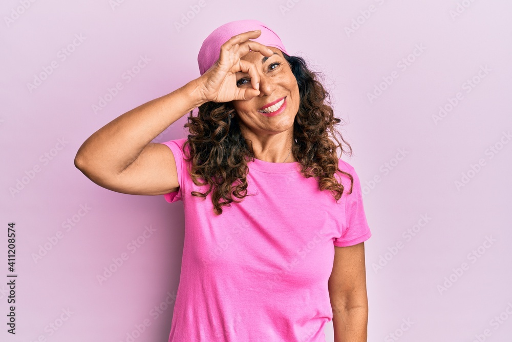 Middle age hispanic woman wearing breast cancer support pink scarf smiling happy doing ok sign with hand on eye looking through fingers Middle age hispanic woman wearing breast cancer support pink scarf smiling happy doing ok sign with hand on eye looking through fingers