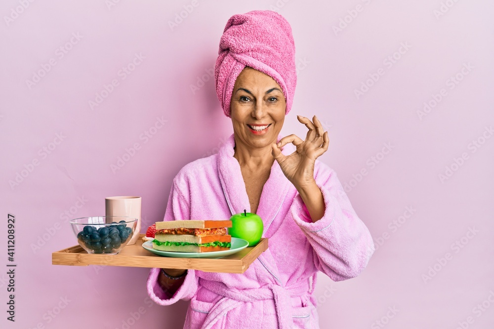Middle age hispanic woman wearing bathrobe holding breakfast tray doing ok sign with fingers, smiling friendly gesturing excellent symbol Middle age hispanic woman wearing bathrobe holding breakfast tray doing ok sign with fingers, smiling friendly gesturing excellent symbol