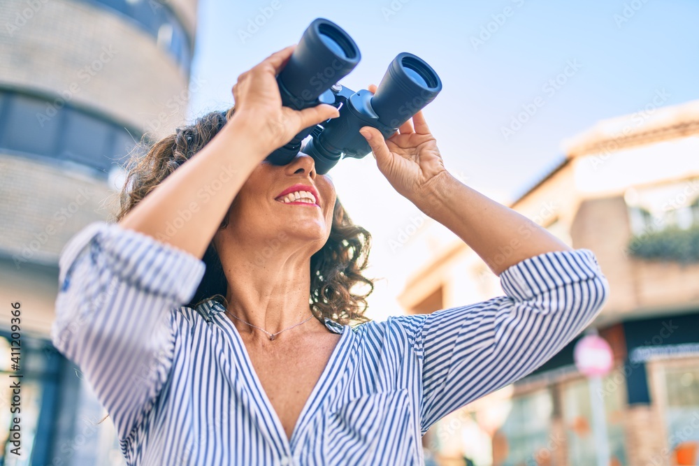 Middle age hispanic woman smiling happy looking for new opportunity using binoculars at the city.