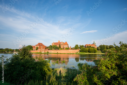 ancient castle of the Teutonic Order of Malbork (Marienburg) on ​​the other side of the river on a clear sunny day against the background of green foliage and blue sky in Poland