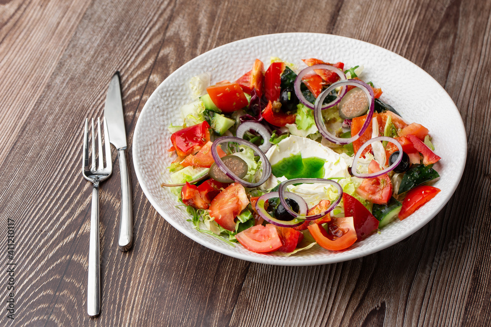 Greek salad with fresh vegetables: tomato, cucumber, red bel pepper, lettuce, onion, olives and cheese. Close-up on a white round plate with cutlery by side on a wooden background. Healthy food. Greek salad with fresh vegetables: tomato, cucumber, red bel pepper, lettuce, onion, olives and cheese. Close-up on a white round plate with cutlery by side on a wooden background. Healthy food.