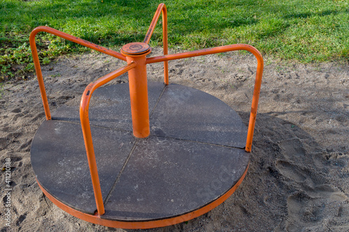 Round orange children carousel with shabby central torsion axis and four handrails with anti-slip coating which divided into sections. Carousel is in sandbox, covered with sand, green lawn behind it.