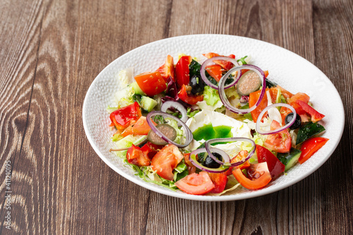 Greek salad with fresh vegetables: tomato, cucumber, red bel pepper, lettuce, onion, olives and cheese. Close-up on a white round plate on a wooden background with a copy space. Healthy food.
