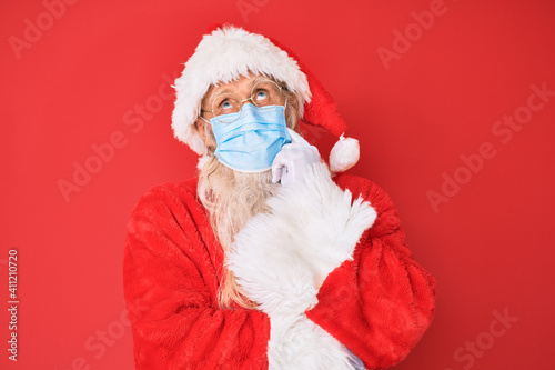 Old senior man wearing santa claus costume wearing safety mask thinking concentrated about doubt with finger on chin and looking up wondering