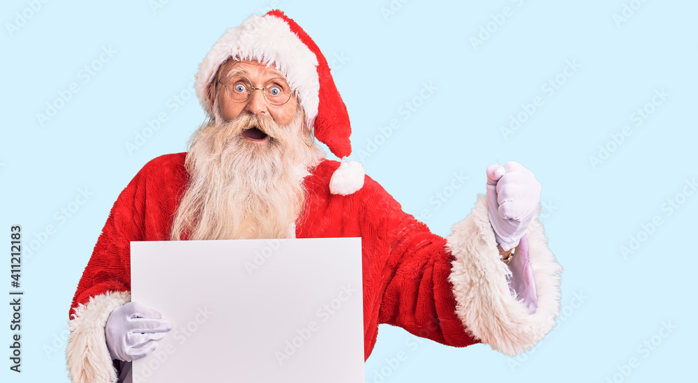Naklejka premium Old senior man with grey hair and long beard wearing santa claus costume holding banner screaming proud, celebrating victory and success very excited with raised arms