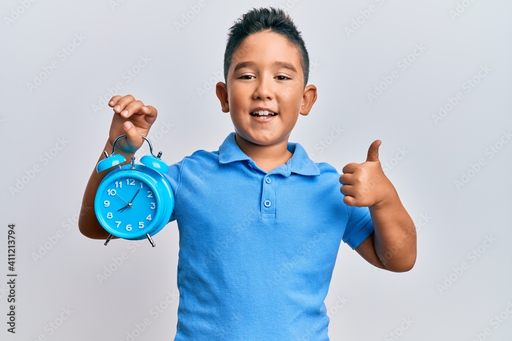 Little boy hispanic kid holding alarm clock smiling happy and positive, thumb up doing excellent and approval sign Little boy hispanic kid holding alarm clock smiling happy and positive, thumb up doing excellent and approval sign