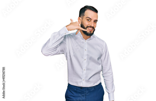 Young man with beard wearing business shirt smiling doing phone gesture with hand and fingers like talking on the telephone. communicating concepts.
